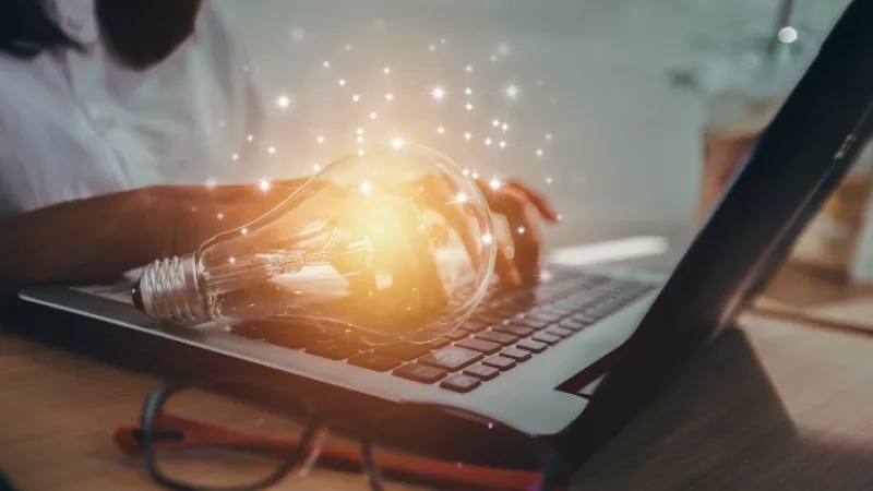 Woman working on laptop with glowing light bulb in hand on desk with glasses in front of laptop.