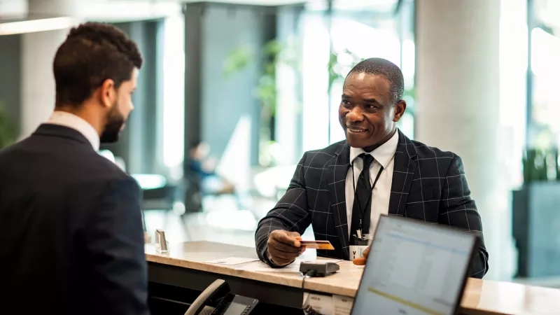 Two men in suits smile at a hotel check-in counter with a laptop and a phone.