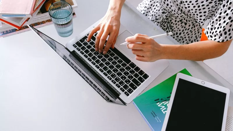 Woman typing on laptop with pen in hand, glass of water, and tablet on desk.
