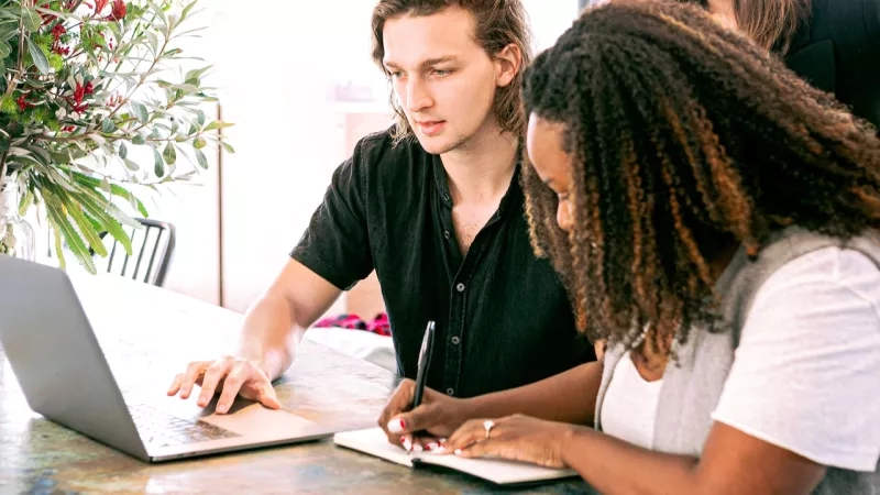 Two people working together on a laptop with a woman writing notes next to them in an office.
