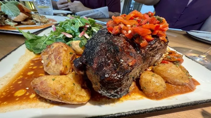 Steak and potatoes on a plate with sauce and peppers, with people and plates in the background.