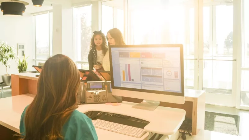 Hotel software on a computer with people in the background and windows with trees outside.