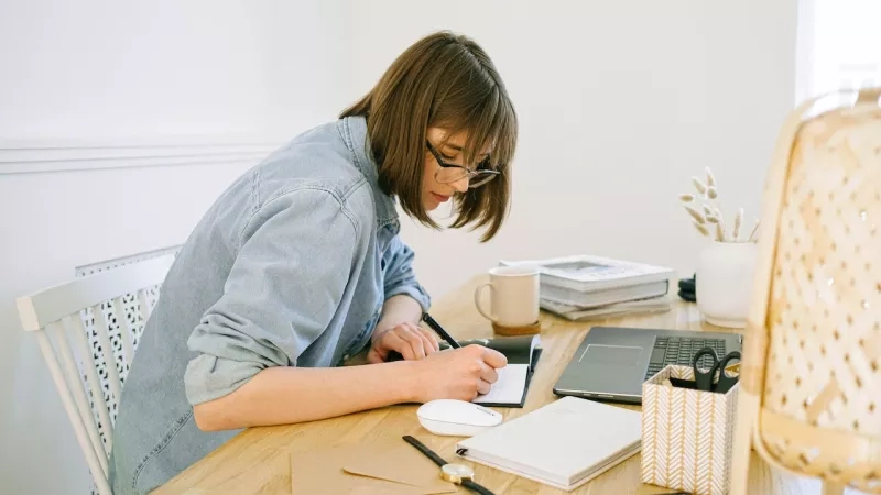 A woman is writing on a notepad at a table with a laptop and other stationery items.