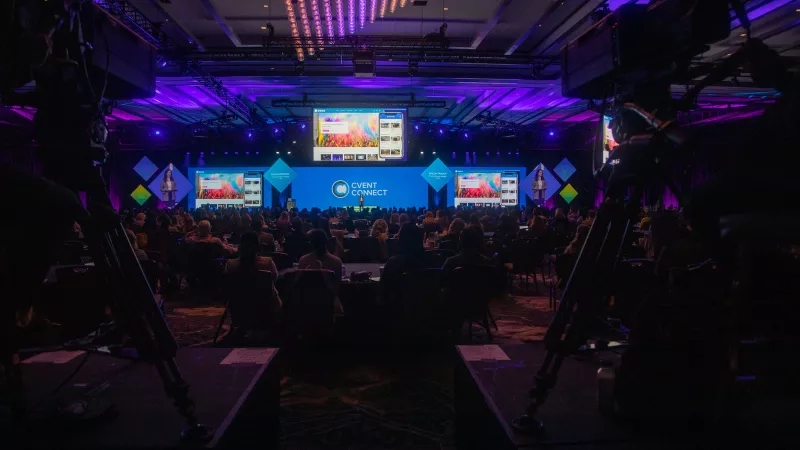 A conference room with a large audience, a stage, and multiple monitors displaying event information.