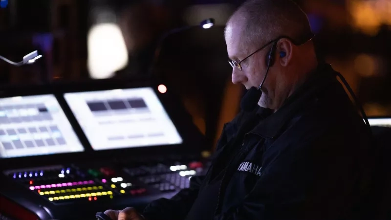 A man is sitting in front of two monitors and a mixing board, working at a sound desk.