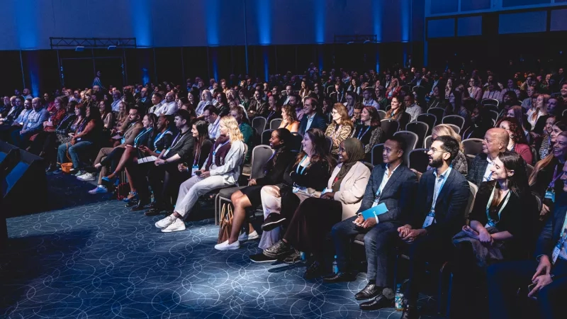 A large group of people are sitting on chairs in an auditorium with blue lighting and a stage.