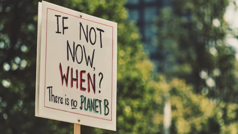 "Sign says 'If not now, when? There is no Planet B' in front of trees and architecture."