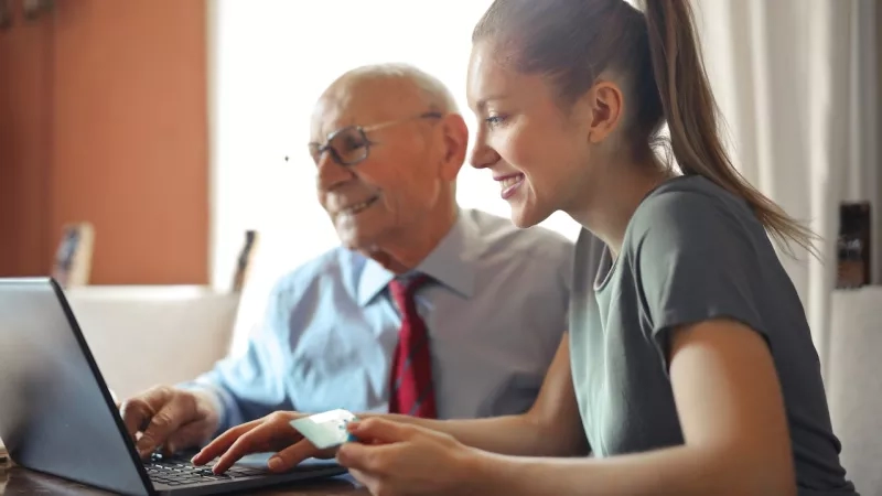 A grandfather and granddaughter use a laptop and credit card together at a table in a home.