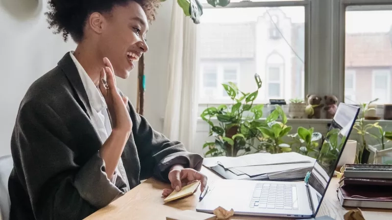 A woman is working on a laptop in an office with a desk and a window.