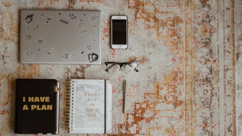 Top view of laptop, iPhone, notebook, pen, and eyeglasses on a rug with text "I have a plan".