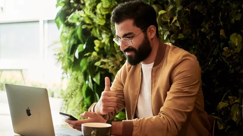 A man is working on a laptop while holding his phone and giving a thumbs up.