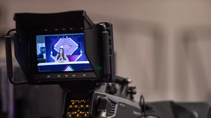 A video camera screen showing a woman in front of a blue and purple background.