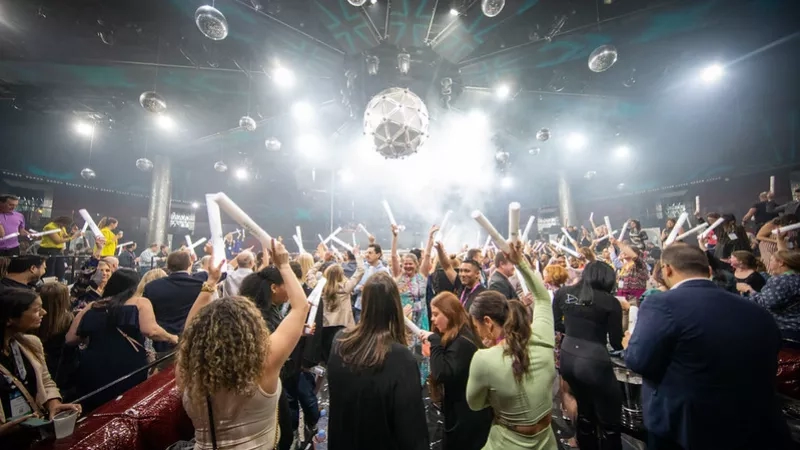 Crowded party room with many people and lights in the ceiling, some are holding up their white cups.