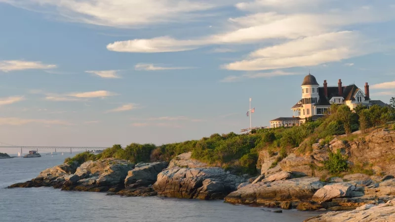 Inn on the water with rocky coast, blue water, bridge, and clouds in the sky.