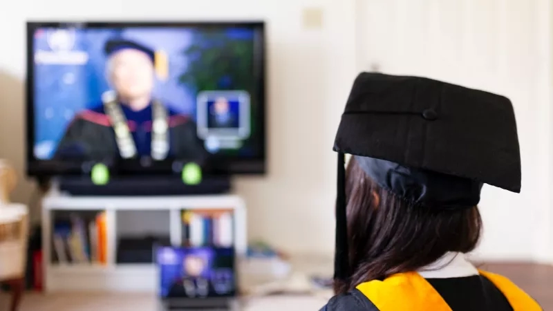 A girl in graduation gown is watching the graduation ceremony on TV at home.