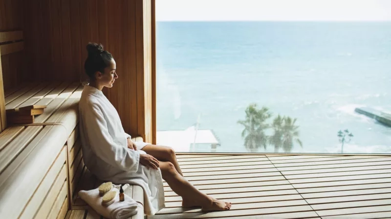 Woman at a wellness hotel spa looking out over the ocean in a wooden sauna.