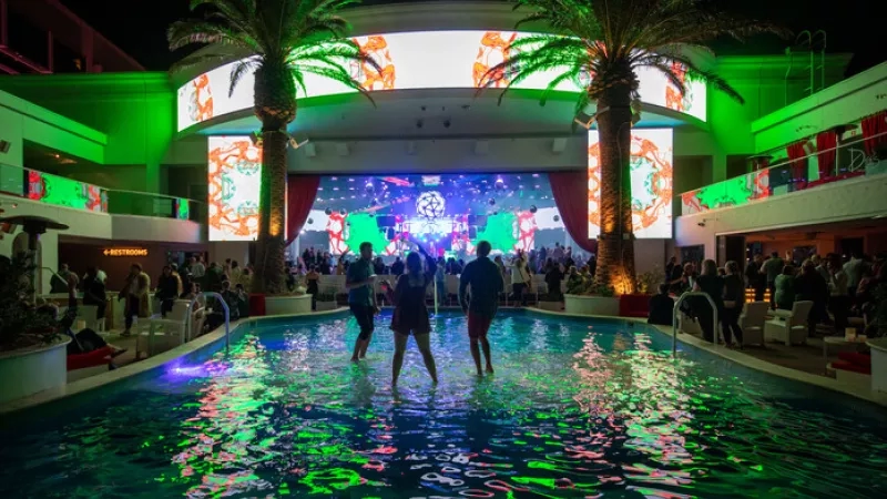 People are standing in a pool at night, illuminated by green lights, with a crowd and stage in the background.