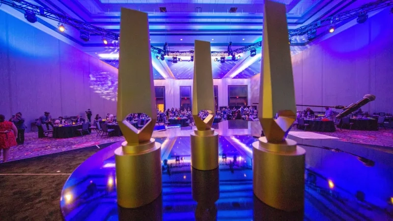 Gold awards stand on a blue table in front of a crowd in a purple lit room.
