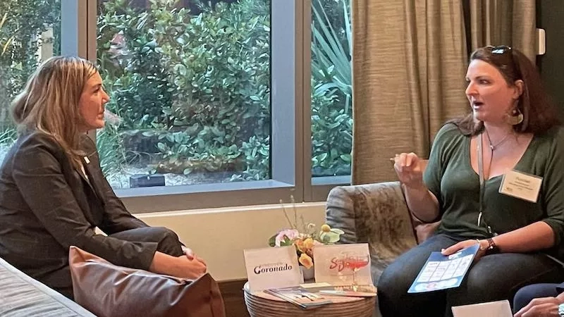 Two women are sitting and talking to each other inside the room in front of the window.