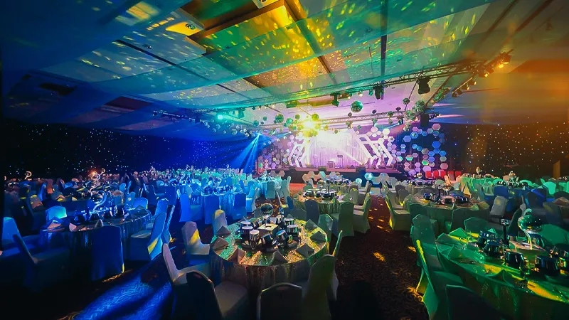 The ballroom is decorated with multicolored lighting and has many tables and chairs in it.