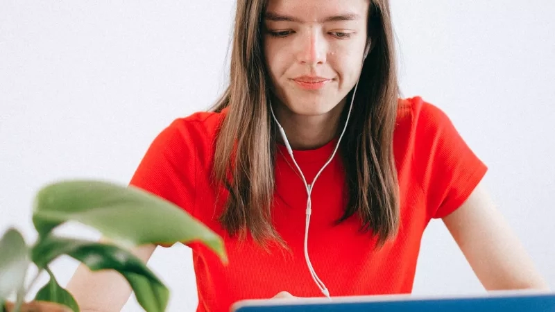A woman in a red shirt with earphones is working on a laptop at her desk.