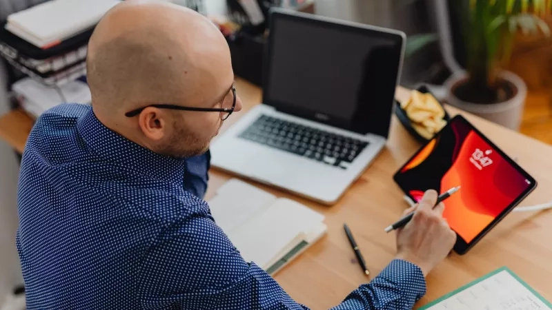 Bald man writing on a tablet while sitting at a desk with a laptop and notebook.