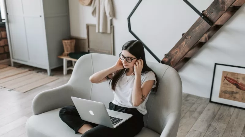 A woman is sitting on a chair with a laptop and headphones in a room with a staircase.