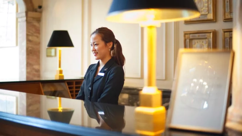 Woman in black blazer standing behind hotel front desk with a smile on her face.