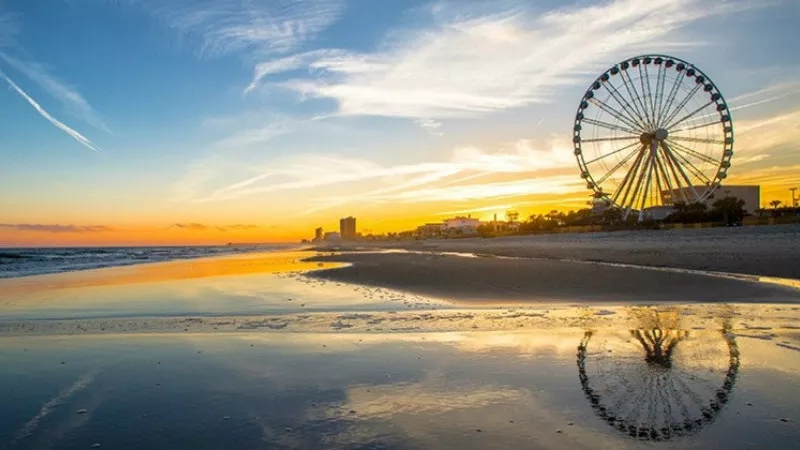 A beach with a Ferris wheel and a sunset sky with clouds and buildings in the distance.