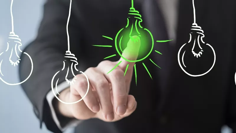 A businessman touches a green light bulb with three light bulbs hanging from the ceiling in the background.