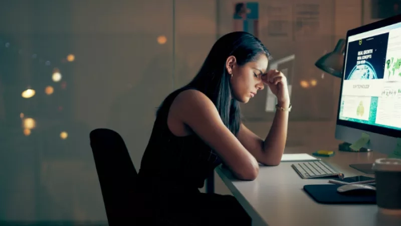 A woman in a black dress is working late at the office and is feeling tired.