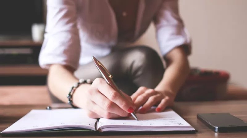 Woman writing in notebook with pen and cell phone on table, blurred background of TV and couch.