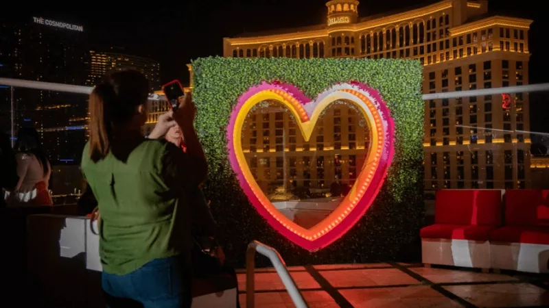 Woman takes a photo of a heart-shaped LED sign with a Bellagio hotel in the background.