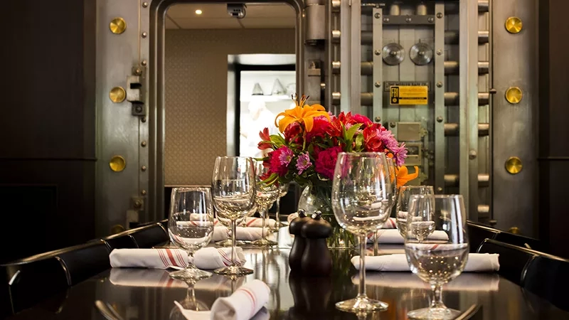 A dining table with wine glasses, napkins, and a flower vase is set in a room with a vault door.