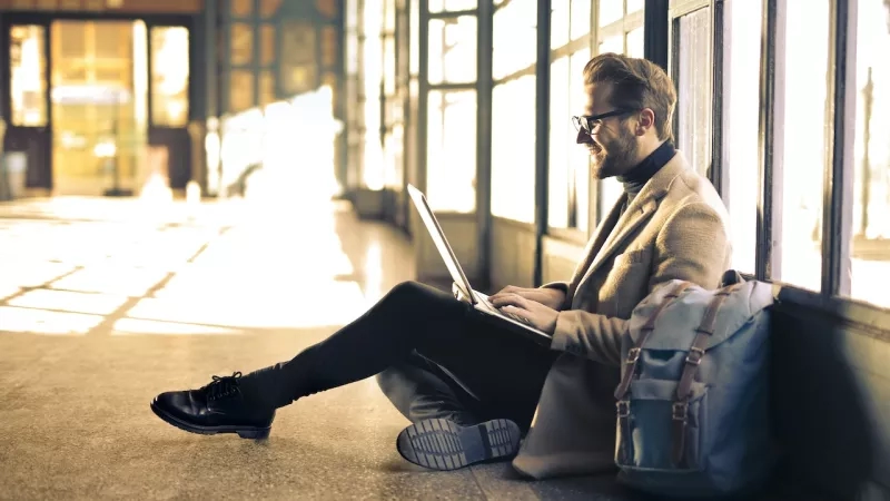 A man using a laptop on a bench with a backpack in a waiting area.