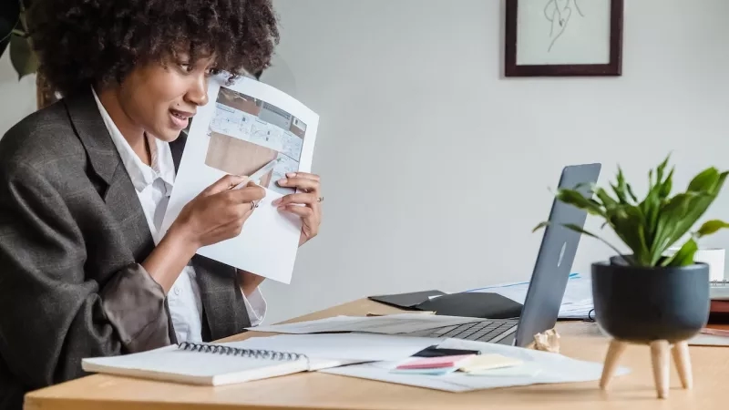 A woman sitting at a desk with a laptop, notebook, and plant, reviewing architectural plans.