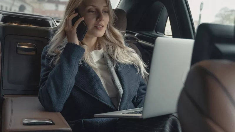 A woman on her phone sitting in a car with a laptop on her lap.