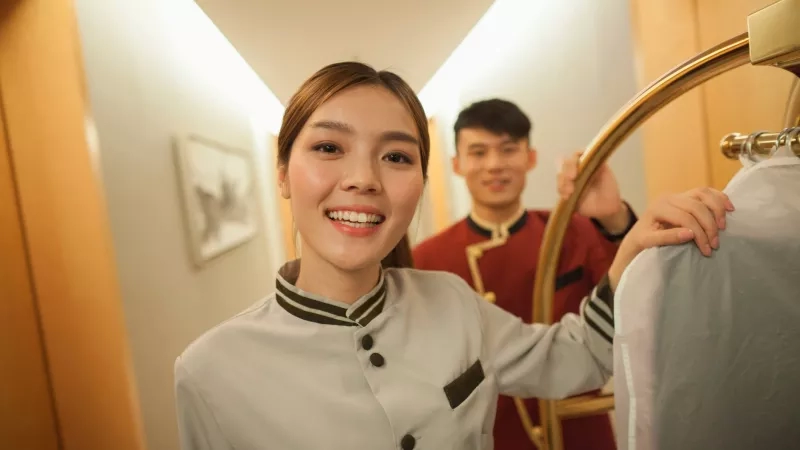 Female hotel staff wearing a uniform and smiling while holding a clothing rack in the corridor.