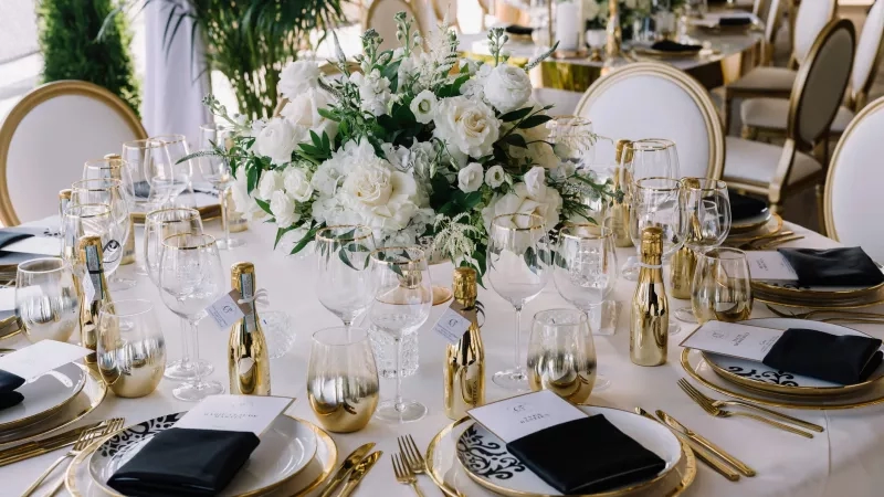 A hotel wedding reception table with white and gold decor, black napkins, and lush greenery.