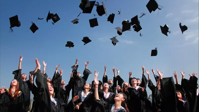 Graduates throw their caps in the air while wearing black graduation gowns and white stoles.