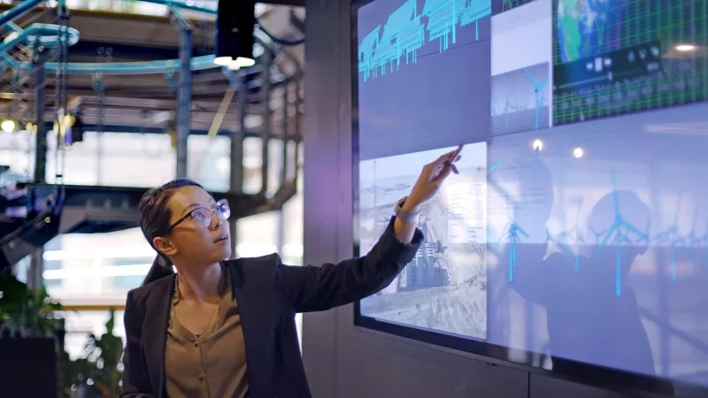 A woman is pointing at a large screen with data, windmills, and a building.