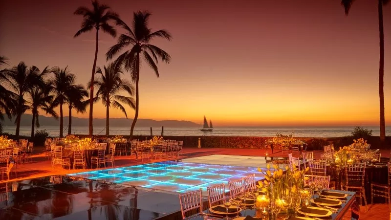 Banquet tables with candles and flowers are set up by the pool in front of the beach at sunset.
