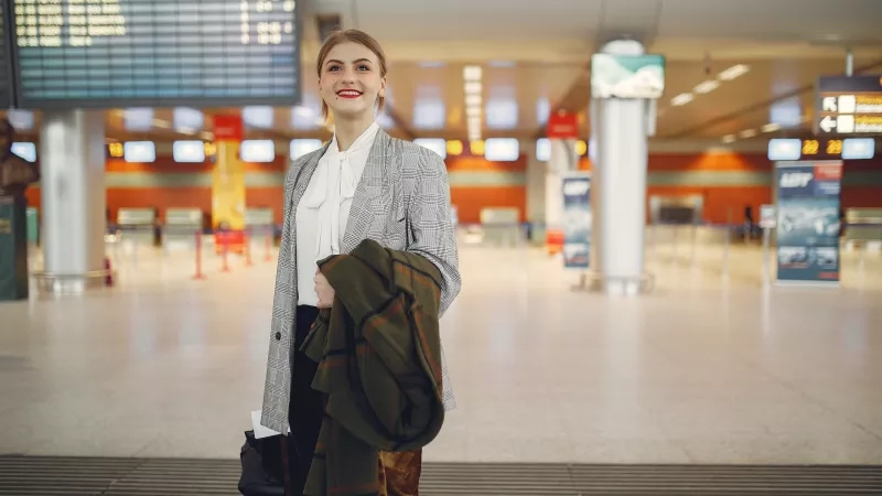 A woman at an airport holding a jacket and a suitcase in one hand and smiling.