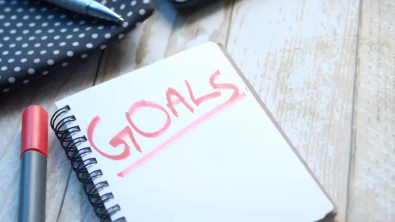 Notebook with the word goals written on it in pink marker on a wooden table with other office supplies.