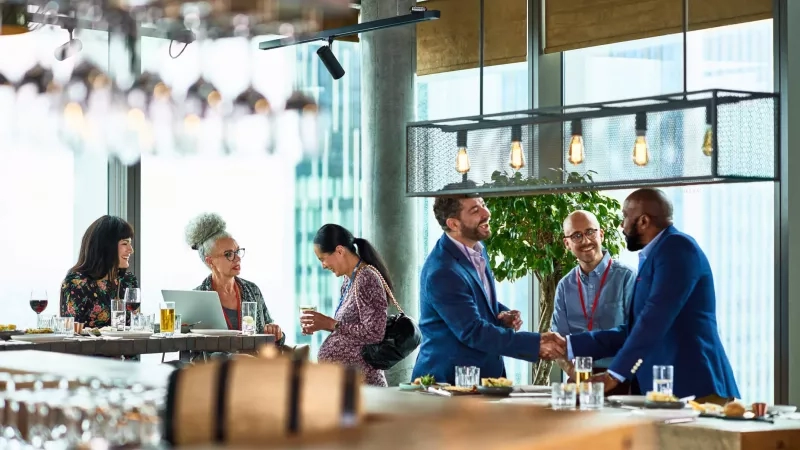 Two men greet each other at a restaurant event in front of a wooden table with drinks and food.
