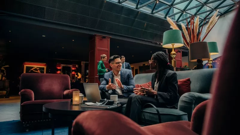 Man and woman talk in a hotel lobby with a laptop on a table between them.