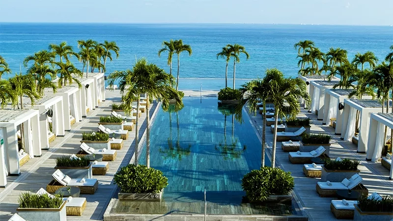 Aerial view of a hotel pool, with ocean views, cabanas, and palm trees.