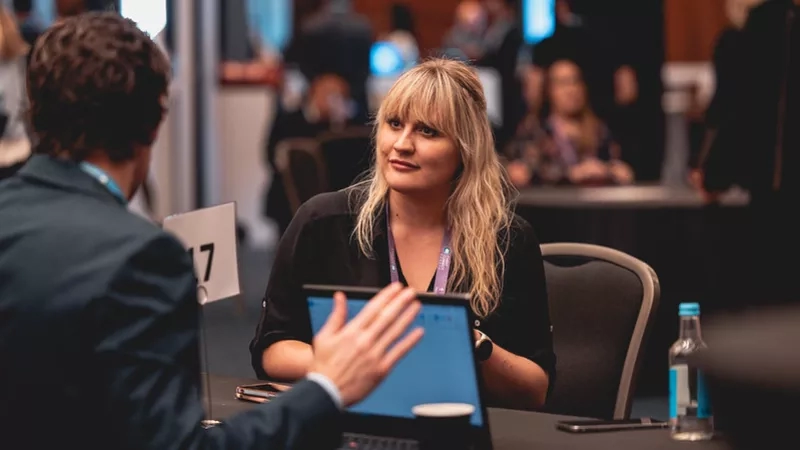 Two people sitting at a table with a laptop and bottle of water at a conference.