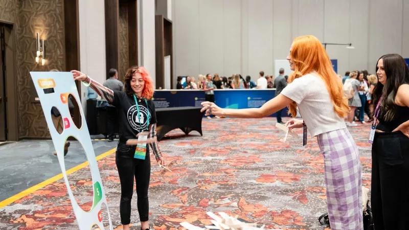 Three women at a tradeshow booth with an interactive game where they throw paper airplanes at a target.