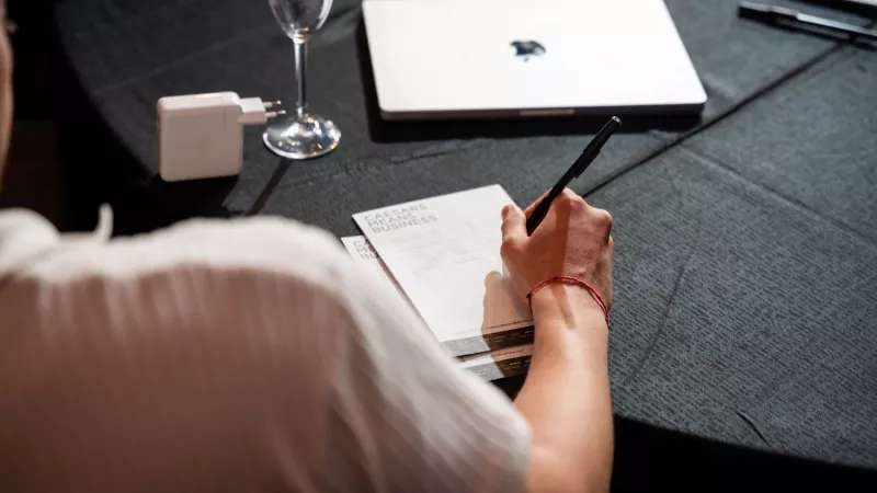 A person with a pen writing on a piece of paper at a table with a laptop and wine glass.